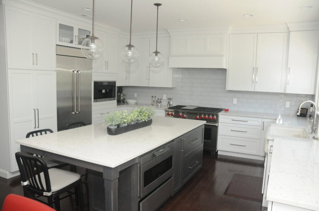 Modern kitchen with white cabinets, stainless steel appliances, a large island with a black base and white countertop, three pendant lights, four chairs, and a rectangular planter with green plants in the center of the island.