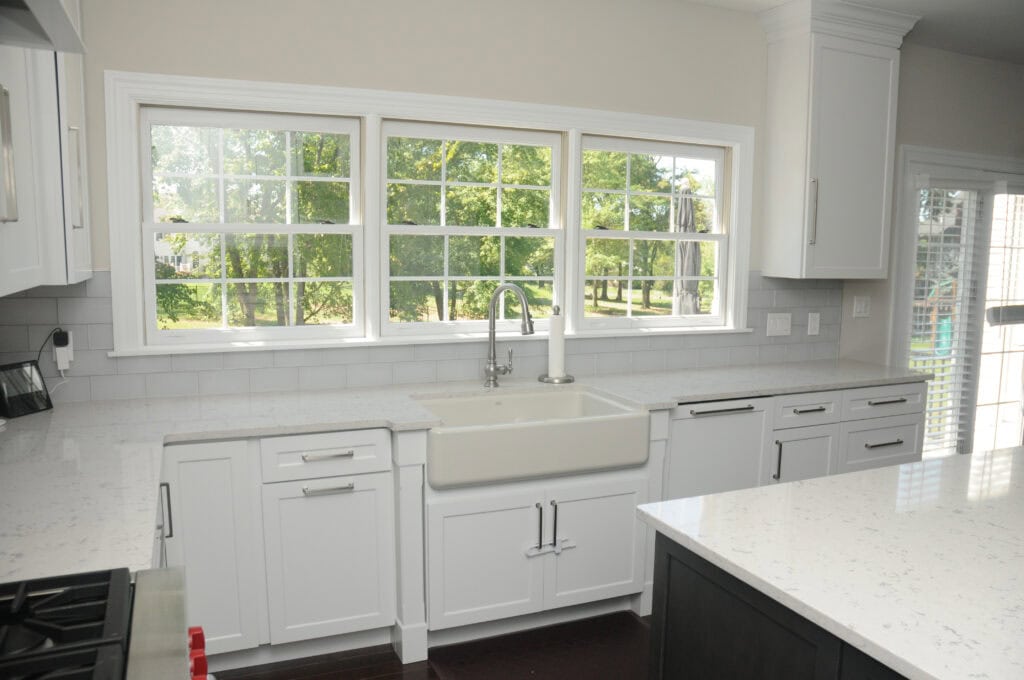 Bright modern kitchen with white cabinets, a farmhouse sink, stainless steel faucet, and large windows overlooking a green, leafy backyard. The countertops are white with subtle gray patterns.