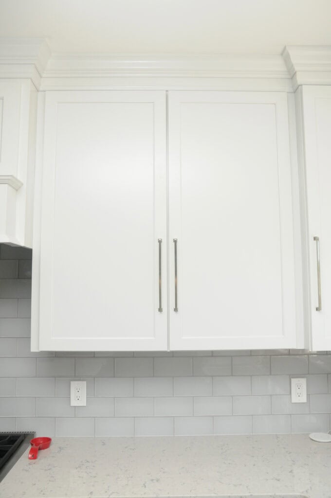 White kitchen cabinets with silver handles above a light gray subway tile backsplash and a white marble countertop. A red measuring cup sits on the counter near an electrical outlet.