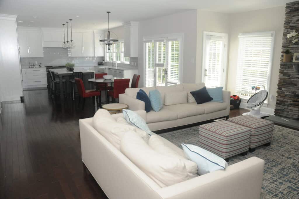 Modern open-concept living room and kitchen with white walls, dark wood floors, two beige sofas, blue and red accent pillows, striped ottomans, a stone fireplace, and a kitchen island with red chairs.