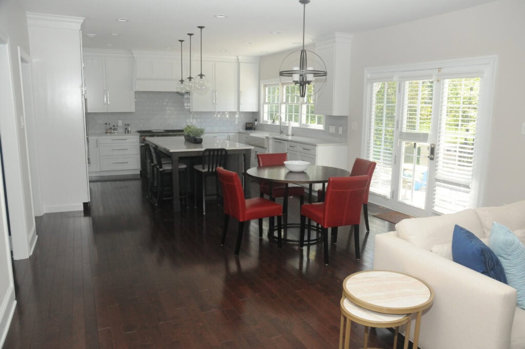 A modern kitchen and dining area with dark wood floors, white cabinets, a black kitchen island with bar stools, a round dining table with red chairs, and large windows letting in natural light.