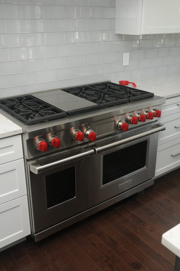 A stainless steel double oven with six gas burners and red knobs is built into white cabinets in a modern kitchen with white subway tile backsplash and dark wood floors.