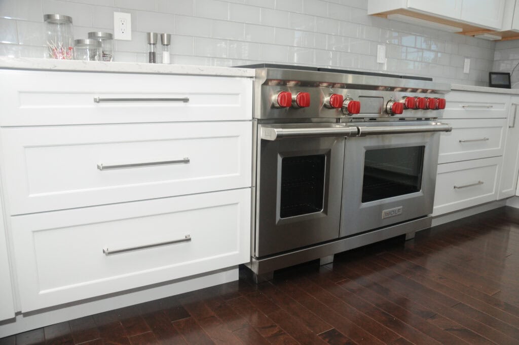 Modern kitchen with white cabinets, stainless steel double oven, red stove knobs, and dark wood flooring. The backsplash is light gray subway tile, and countertops are white.