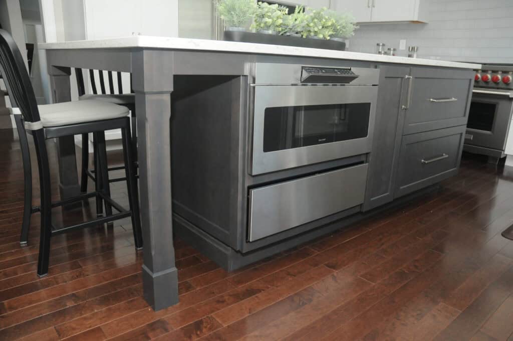 A modern kitchen island with dark cabinets, stainless steel microwave, and drawer, set on polished wood floors. A black chair with a white cushion is placed at the island.