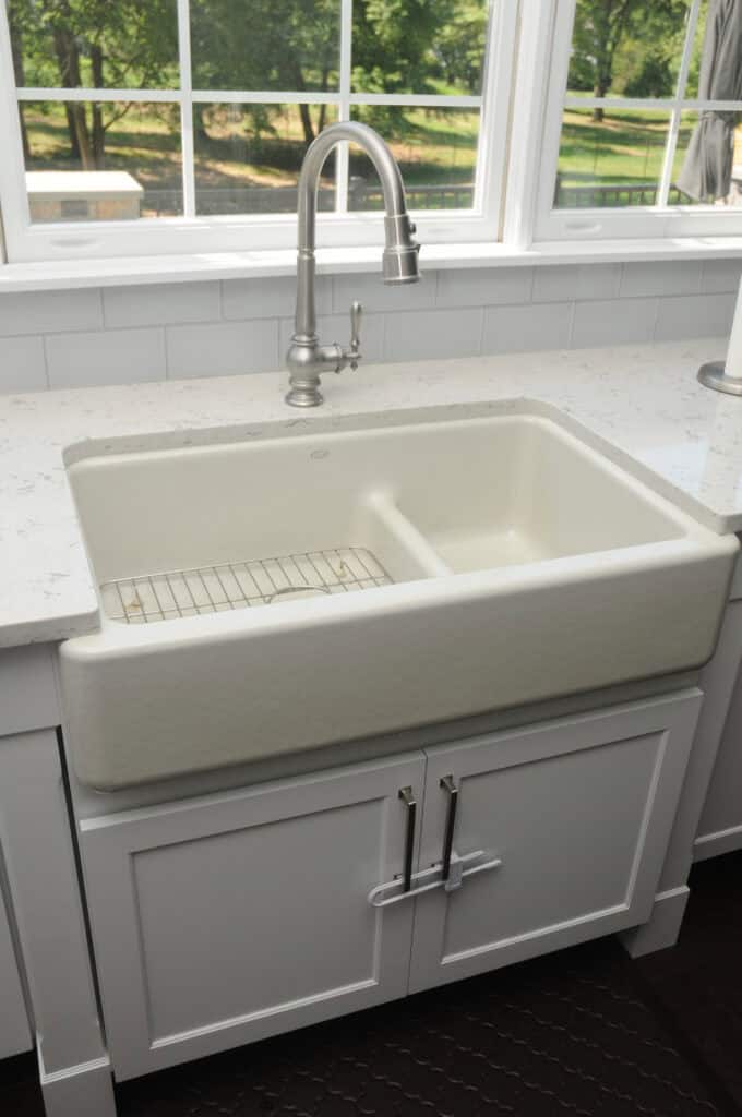 A white farmhouse kitchen sink with a divider and a metal grid insert sits beneath a large window, surrounded by white cabinets and countertops. A modern, brushed metal faucet is installed above the sink.