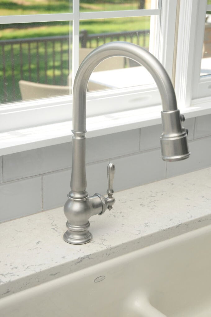 A brushed nickel kitchen faucet with a curved spout and single lever handle is installed on a light-colored countertop in front of a window with a view of a green outdoor area.