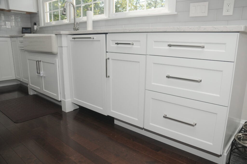 Modern kitchen with white cabinets, stainless steel handles, dark wood flooring, farmhouse sink, and a window above the counter letting in natural light. A brown mat is placed on the floor near the sink.