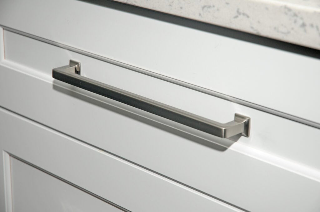 Close-up of a modern silver drawer handle on a white kitchen cabinet with a light-colored, speckled countertop above.
