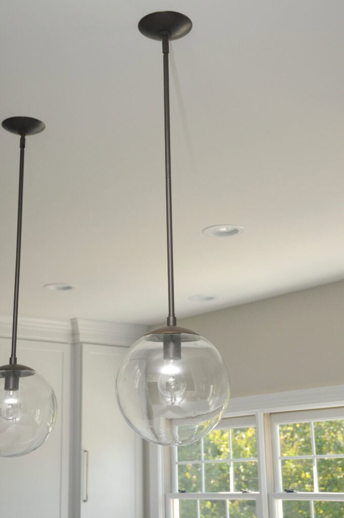 A modern pendant light with a clear glass globe shade hangs from the ceiling next to another identical fixture in a bright room with white walls and large windows.
