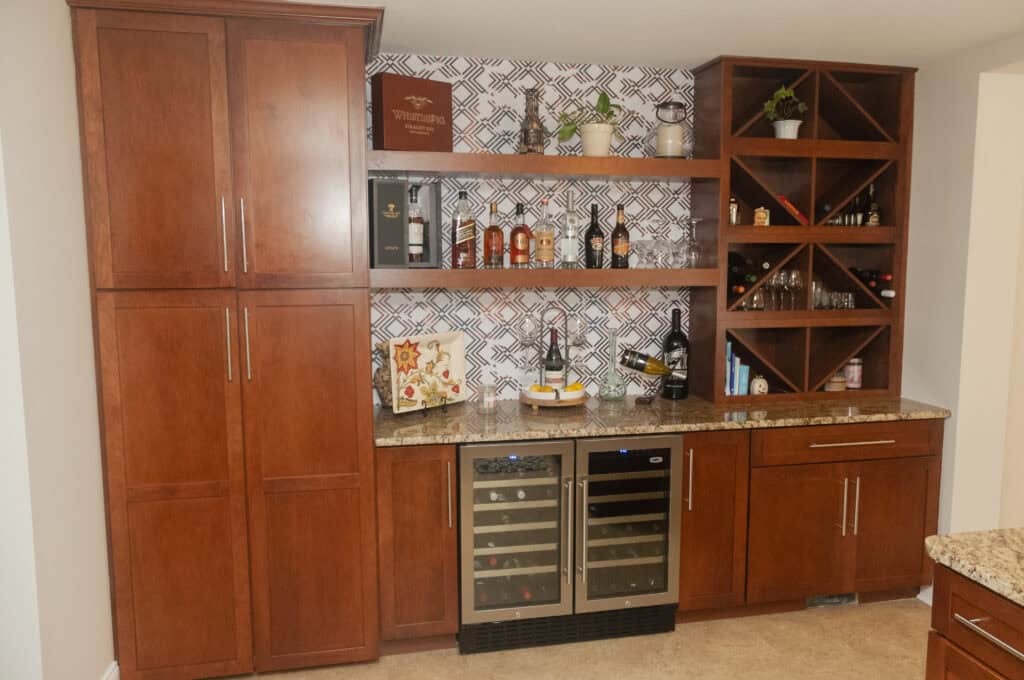 A home bar with wooden cabinets, wine racks, a granite countertop, a double wine fridge, and open shelves holding bottles, glasses, books, and small potted plants. The backsplash features a geometric tile pattern.