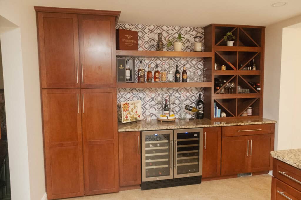 A home bar with wooden cabinets, built-in wine racks, glass-front mini fridges, open shelves with bottles, glasses, and plants, set against a patterned tile backsplash.