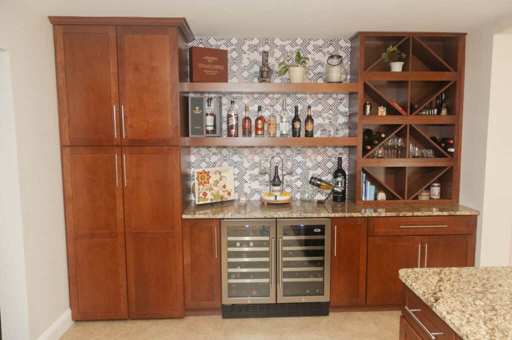 A home bar area with wooden cabinets, two wine coolers, granite countertop, open shelves holding bottles, plants, and decor, and a geometric patterned backsplash.
