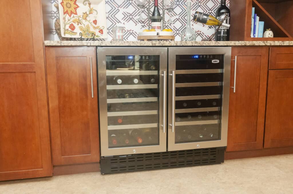 A built-in dual-zone wine cooler with glass doors is installed in a kitchen cabinet with brown wood cabinetry. The wine cooler is stocked with several bottles of wine and has digital display panels on top.