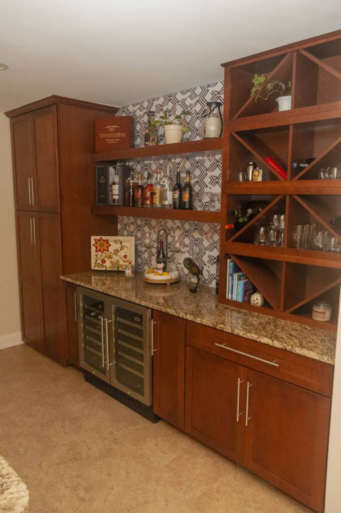 A home bar area with wooden cabinets, granite countertop, a wine rack, a small wine fridge, assorted bottles, glasses, plants, and decorative items against a patterned backsplash wall.