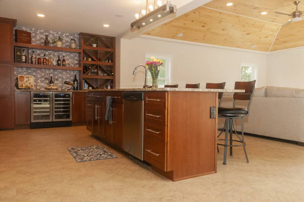 Spacious kitchen with wooden cabinets, an island with a sink, bar stools, wine fridges, open shelves with bottles, geometric backsplash, and a living area with a gray couch and wood-paneled ceiling.