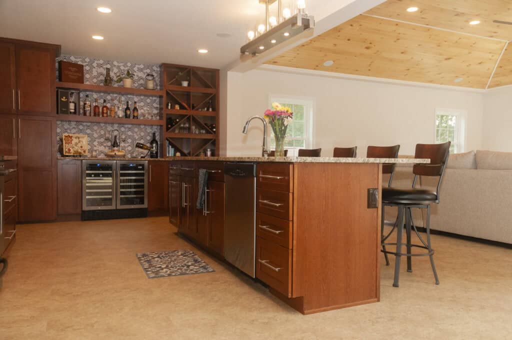 Modern kitchen with wooden cabinets, an island with a sink, bar stools, built-in wine fridge, patterned backsplash, and open shelves with bottles and glasses. Vase with flowers sits on the island. Couch visible in background.