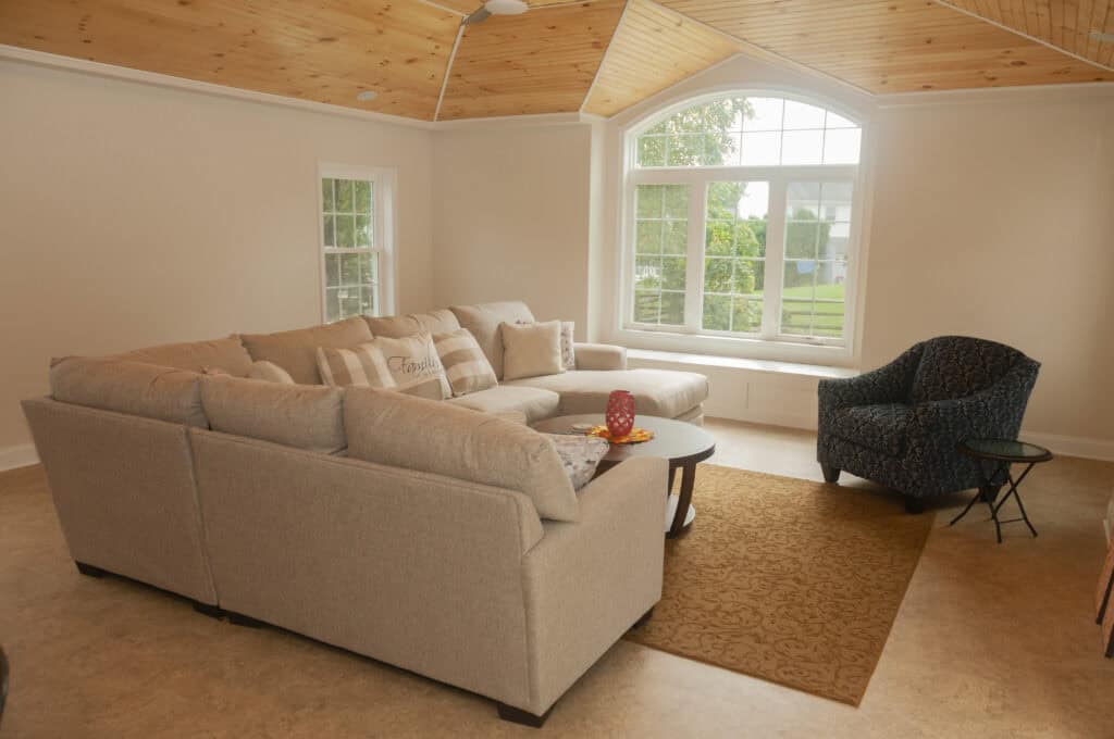 A cozy living room with a large beige sectional sofa, a patterned dark armchair, a small round table, and a tan rug. Large windows let in natural light, and the ceiling is wood-paneled.