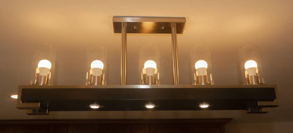 A modern rectangular light fixture with five illuminated light bulbs, each inside a clear glass cylinder, is mounted on a ceiling. Three round recessed lights are installed along the fixtures base.