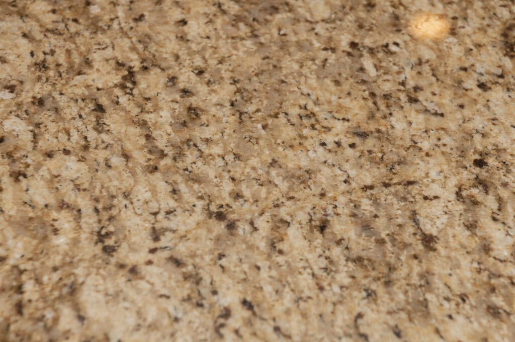 A close-up of a beige granite countertop surface with brown, black, and gray speckles and natural stone patterns. The texture appears smooth and polished, with a small light reflection in the upper right corner.