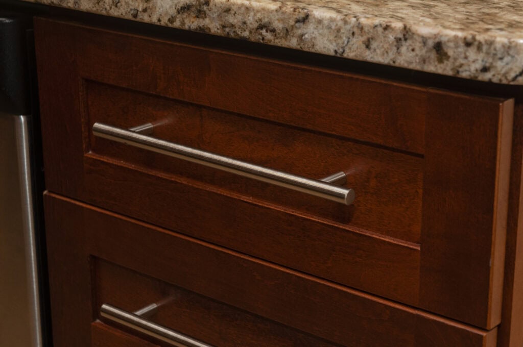 Close-up of a kitchen cabinet with a wooden finish, stainless steel handle, and a granite countertop above.