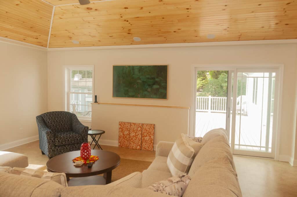 A bright living room with a beige sectional sofa, patterned armchair, round wooden coffee table, wall art, and large sliding glass doors leading to a sunny deck. The ceiling is wood-paneled with recessed lighting.