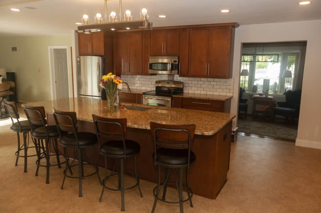 A modern kitchen with wooden cabinets, a large granite island with five barstools, stainless steel appliances, white subway tile backsplash, and a vase of flowers on the counter. A bright dining area is visible in the background.