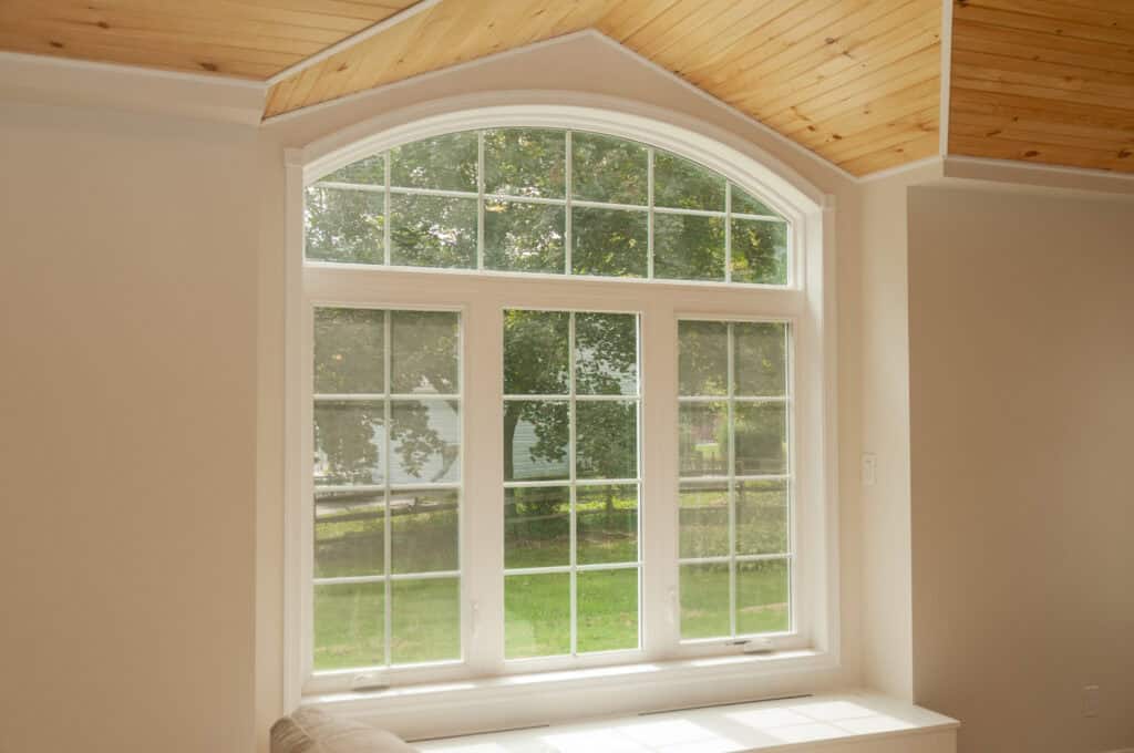 A large arched window with white trim lets in natural light and offers a view of green trees and grass outside. The room features cream-colored walls and a wooden, angled ceiling.