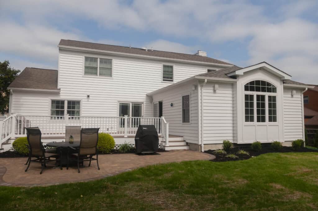 A white two-story house with large windows and a deck, featuring patio furniture and a grill, overlooks a lawn and garden under a partly cloudy sky.