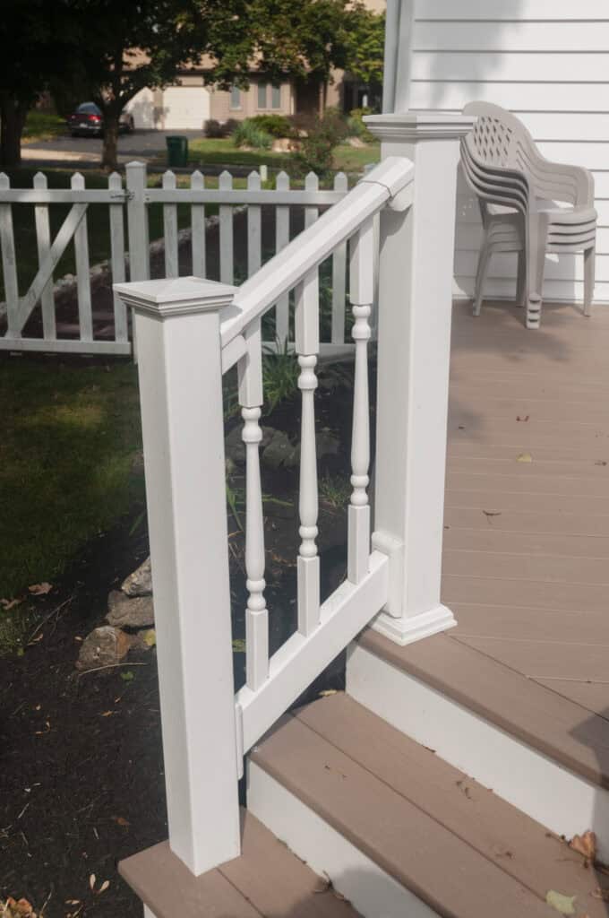 A white wooden handrail with spindles lines the edge of outdoor steps leading to a porch. Stacked patio chairs sit on the porch, and a white picket fence runs along the grassy yard in the background.