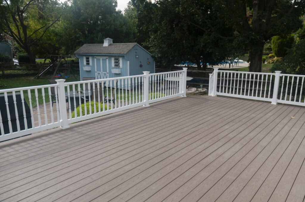 A spacious wooden deck with white railings overlooks a backyard with green grass, trees, and a small blue shed.