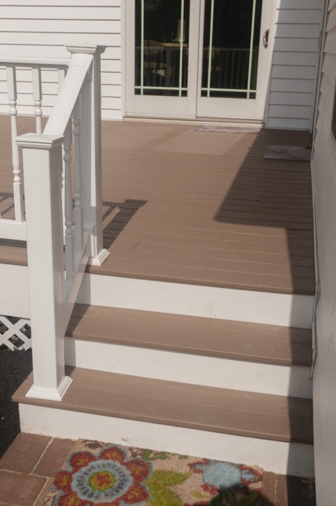 Three wooden steps with a white railing lead up to a small porch outside glass double doors. A colorful floral doormat sits at the bottom of the steps, and sunlight casts shadows on the porch.