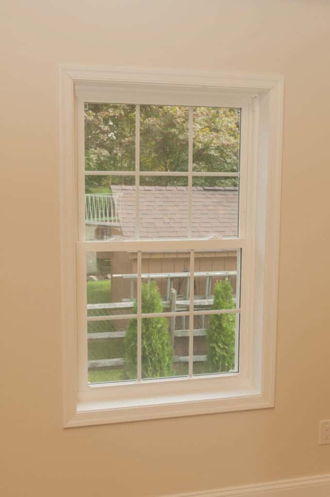 A single window with white trim set in a beige wall, looking out onto a small brown shed, green trees, and a patch of grass in a backyard.