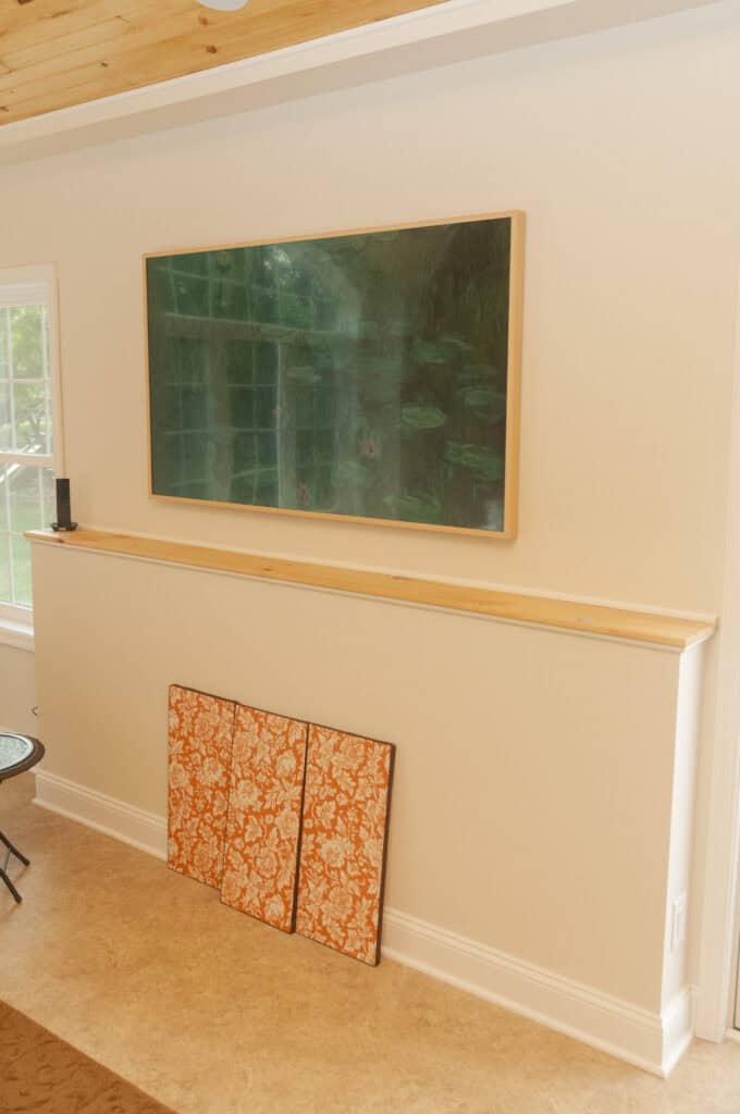 A large framed painting hangs on a beige wall above a narrow wooden shelf. Below the shelf, two orange patterned panels rest against the wall. A window lets in natural light.