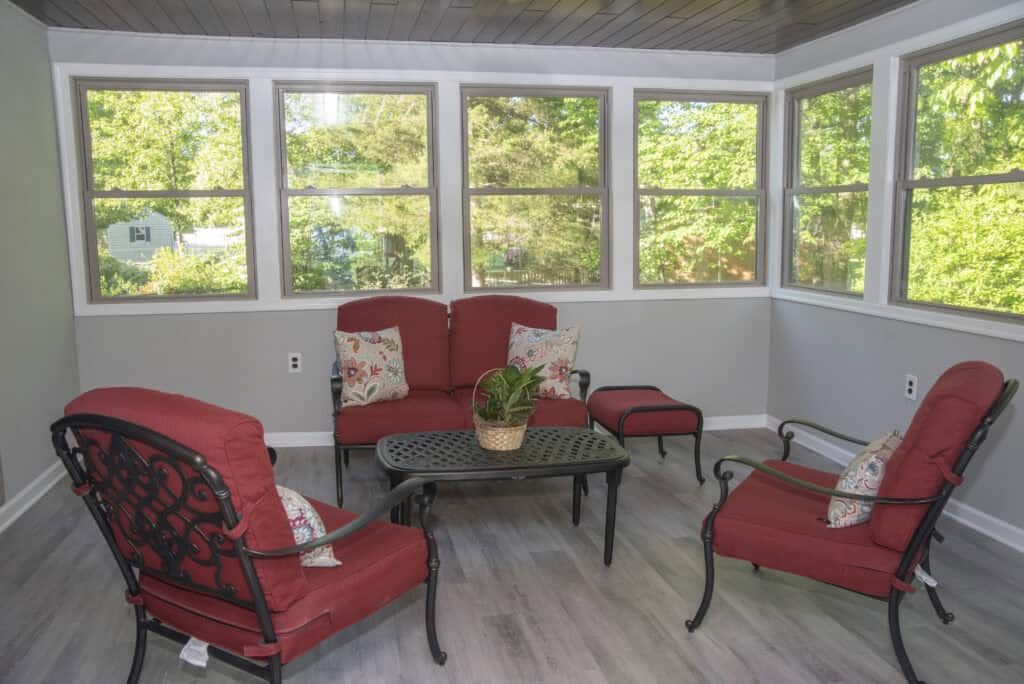 A sunroom with large windows, featuring a black metal patio set with red cushions, a glass-top coffee table, and a potted plant centerpiece. The room has light gray walls and wood-look flooring.