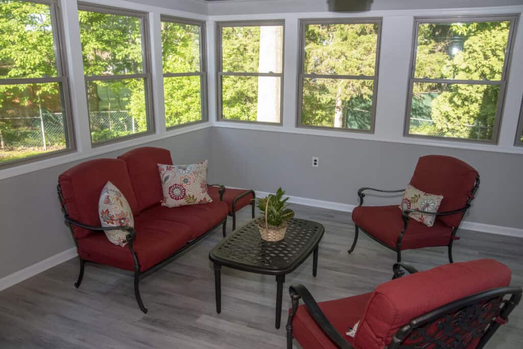 Bright sunroom with large windows, featuring red cushioned chairs, a loveseat with patterned pillows, a black coffee table, and a small potted plant; light gray walls and wood-style flooring. Lush greenery visible outside.