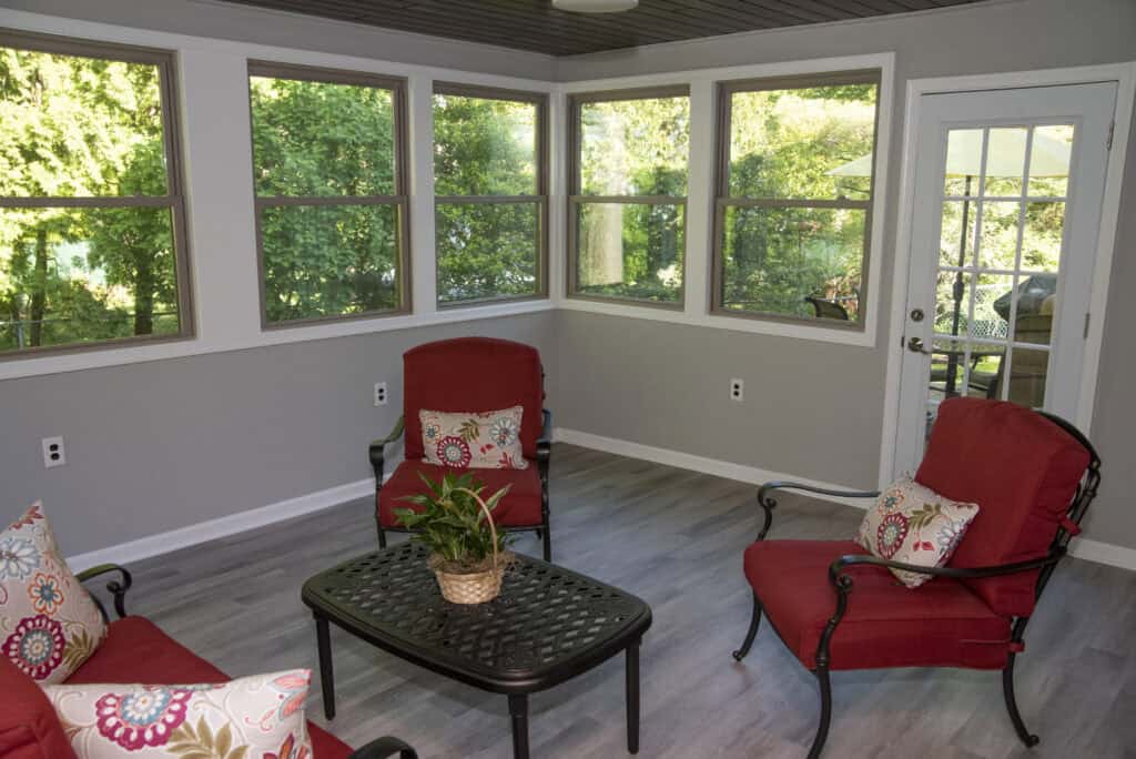 A bright sunroom with large windows, three red cushioned chairs, a black coffee table with a potted plant, and light gray wood flooring. Green trees and a patio umbrella are visible outside.