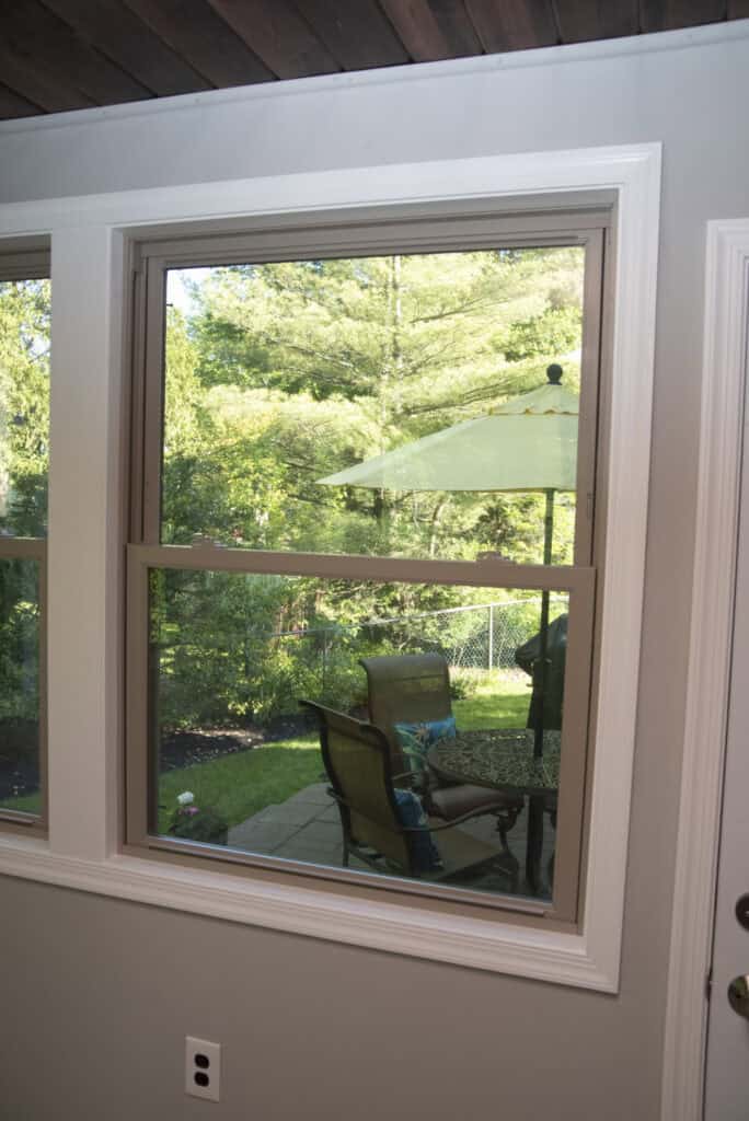 A view through a window from inside a house shows patio chairs and a table with an umbrella outside, surrounded by greenery and trees under daylight.