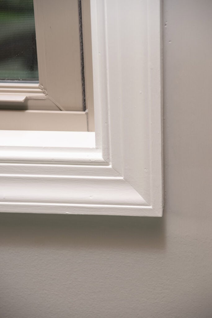 Close-up of a white-framed window on a light-colored wall, showing detailed trim and a small section of the window pane in the top left corner.
