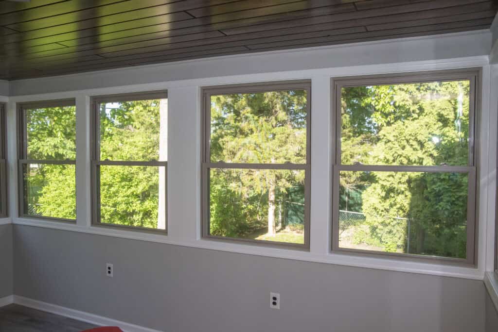 Four large windows in a bright room with light gray walls and a dark wood ceiling, showing a view of green trees and sunlight outside. Two electrical outlets are visible on the wall below the windows.