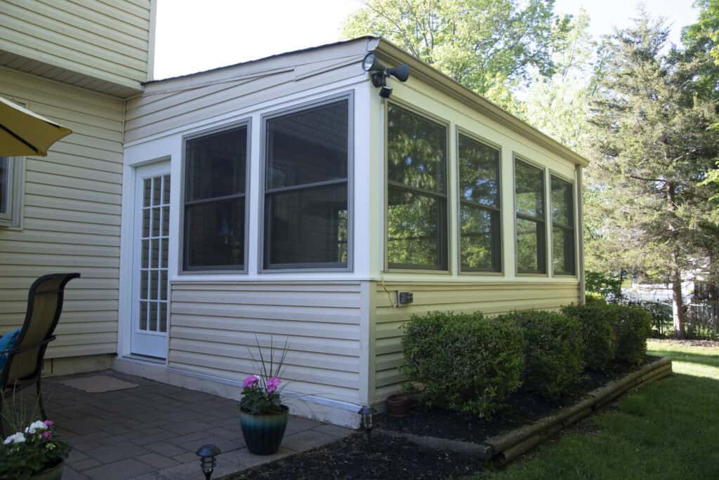 A sunroom with large windows and beige siding attached to a house, surrounded by green bushes and potted flowers, with a patio area and outdoor furniture nearby.