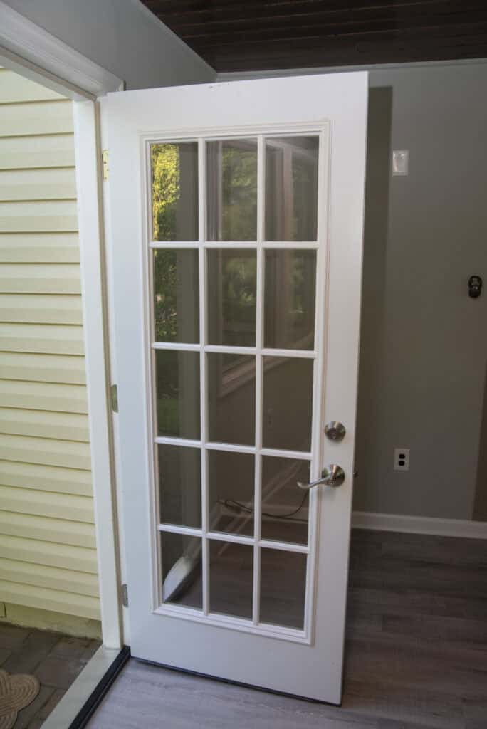 A white door with glass panes divided into a grid is open to the outside. The door has silver handles and is set in a room with gray walls and light wood flooring. Yellow siding is visible outside.