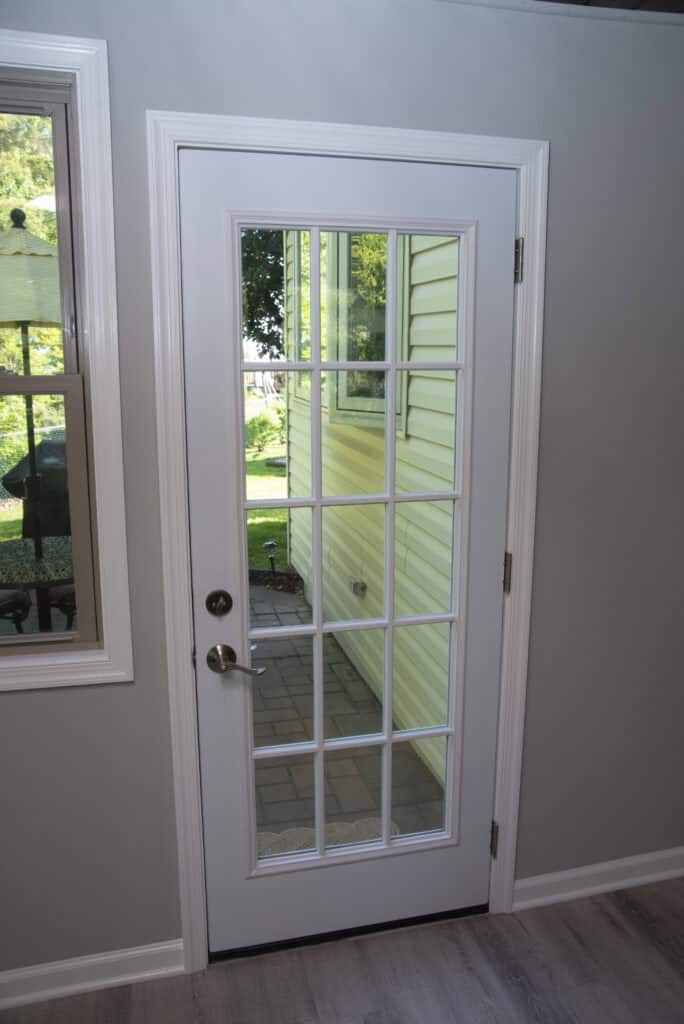 A white-framed glass door with multiple panes leads outside to a patio area, with green grass and a yellow-sided house visible through the glass. A window is on the left, and the interior wall is light gray.