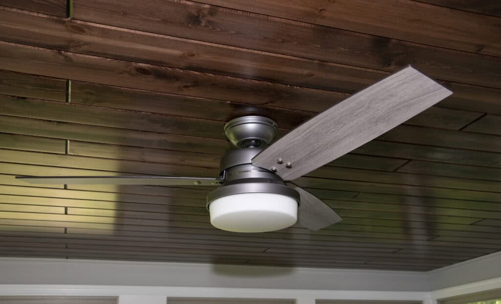 A modern ceiling fan with three blades and a central light fixture is mounted on a wooden plank ceiling. The fan has a metallic finish and the blades are a light gray color.