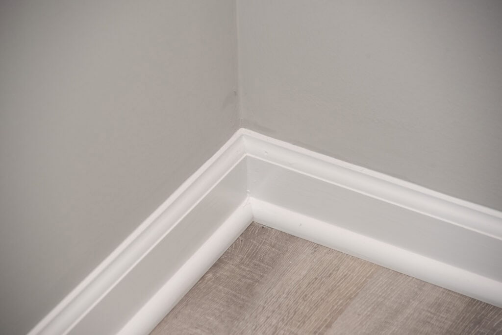 Close-up of the corner of a room, showing light gray walls, white baseboards, and light wood-colored flooring.