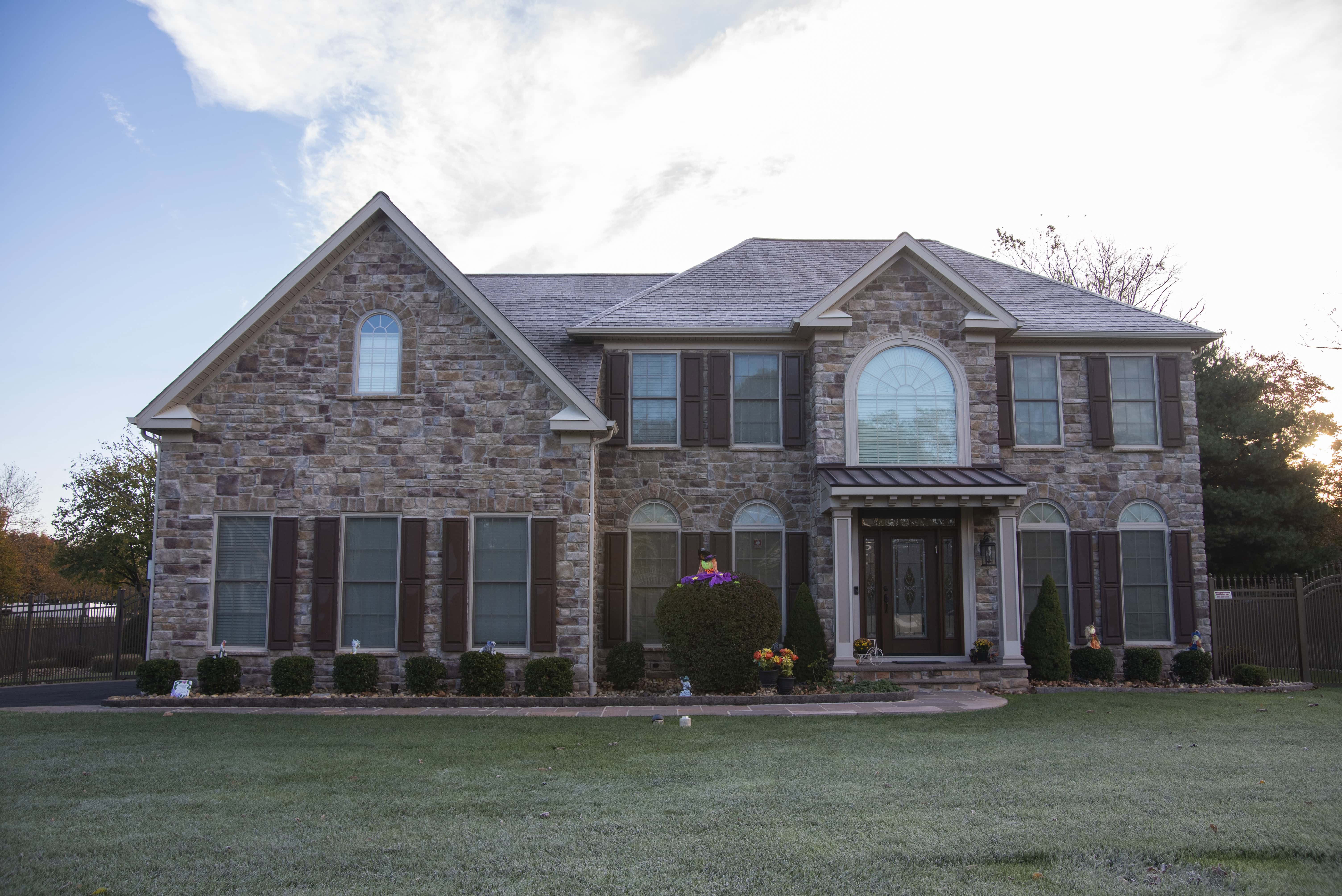 A two-story house with a stone exterior, brown shutters, and a large arched window above the entrance, set against a clear sky with green grass and landscaping in front.