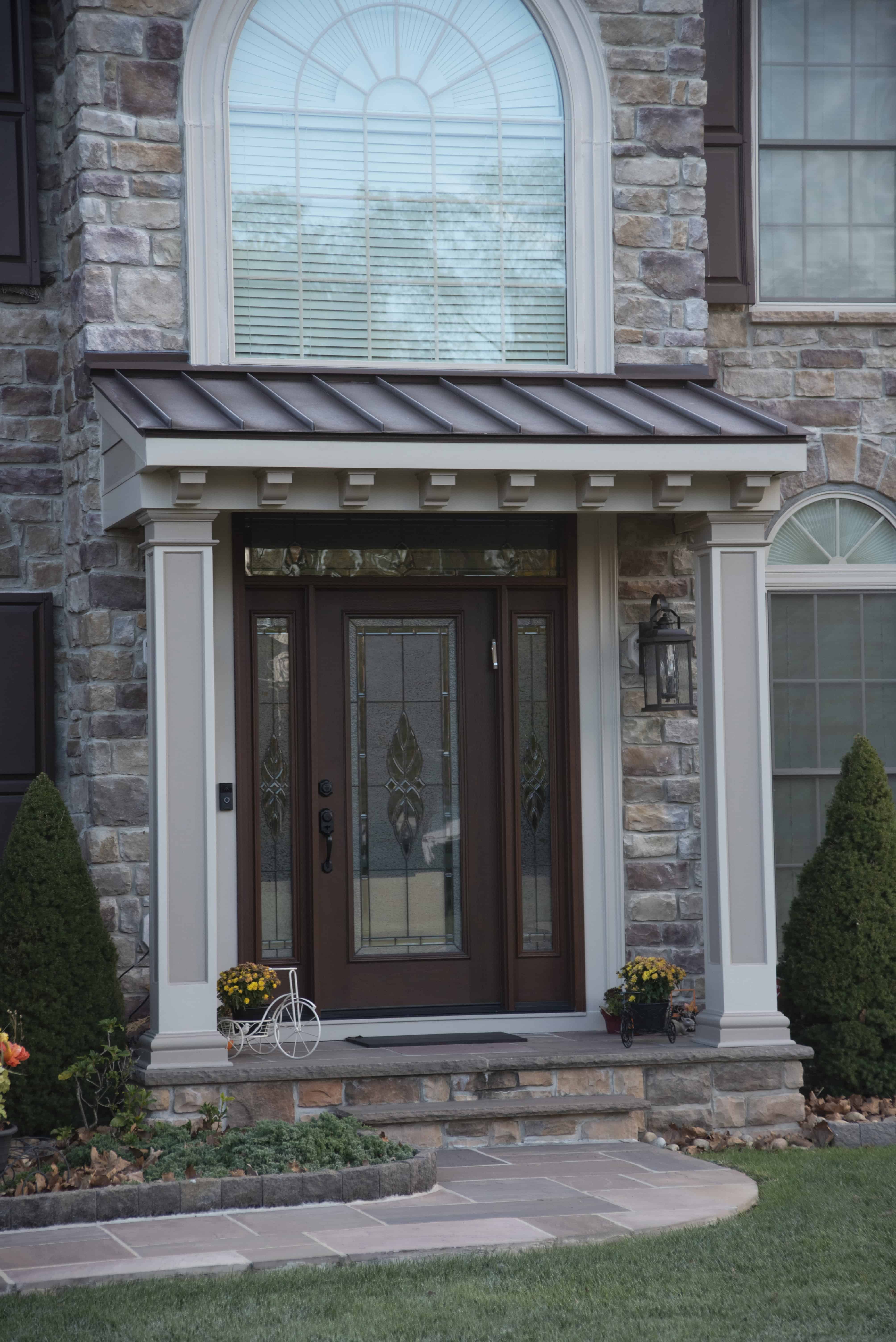 A front door with decorative glass panels, framed by white columns and a metal awning, set in a stone exterior. Potted flowers and a small metal bicycle planter sit on the porch steps. Shrubs and windows surround the entryway.