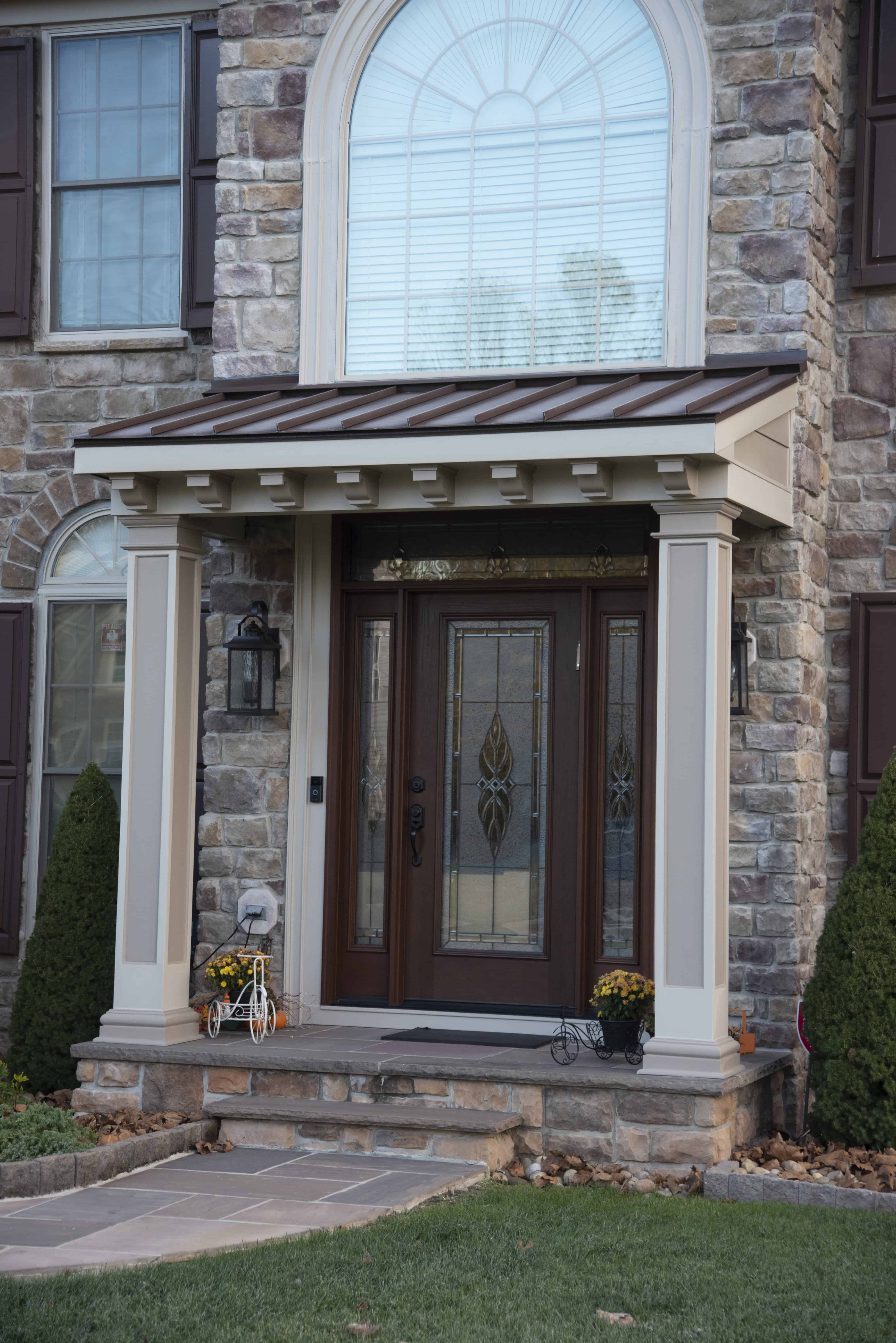 A brown wooden front door with decorative glass panels is set in a stone facade, framed by columns and a small roof. Two potted yellow flowers and a small toy bike decorate the front steps.