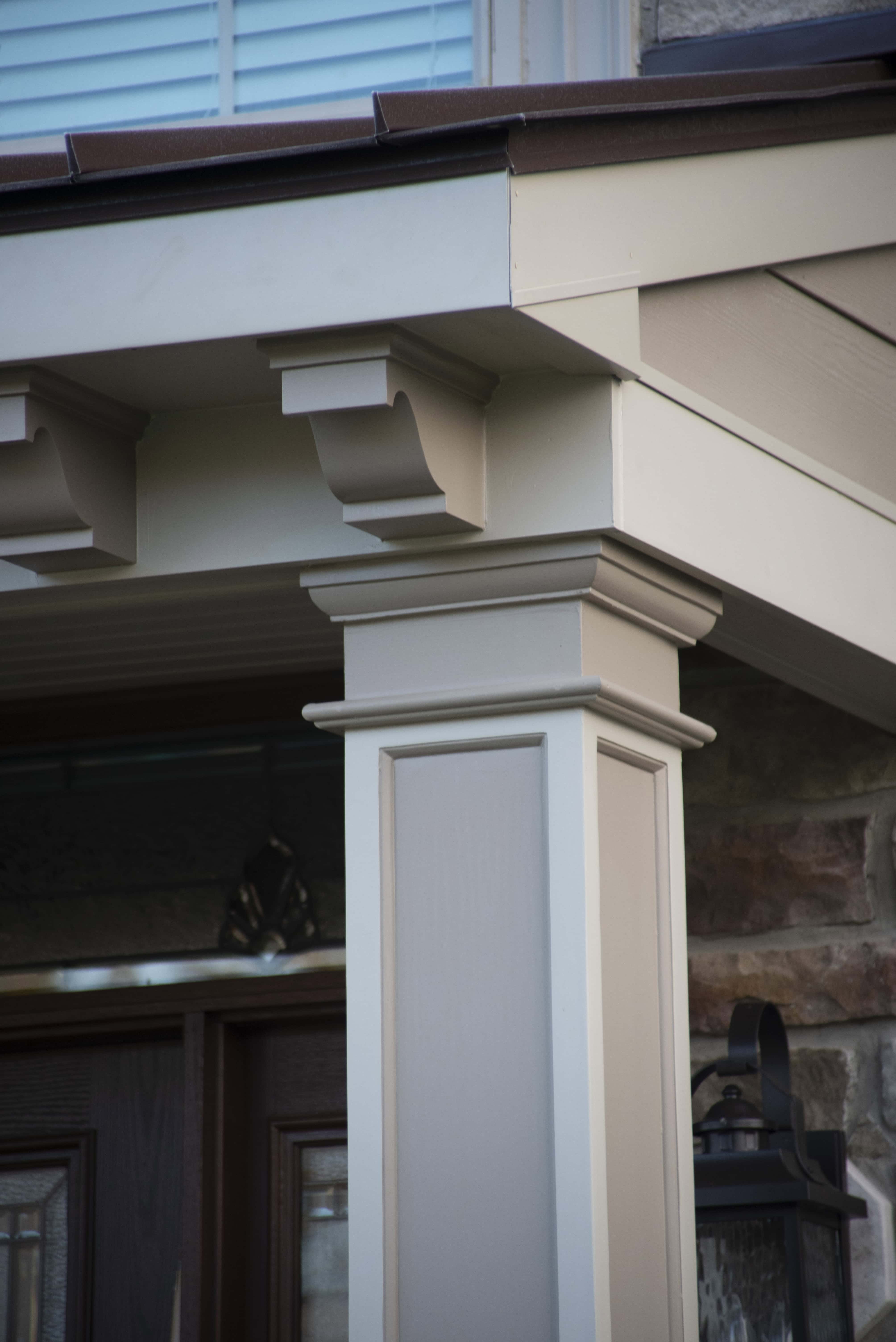 Close-up of a beige porch column with decorative trim supporting a roof overhang, next to a stone wall and wooden door with a glass panel. A black outdoor lantern is mounted on the wall.