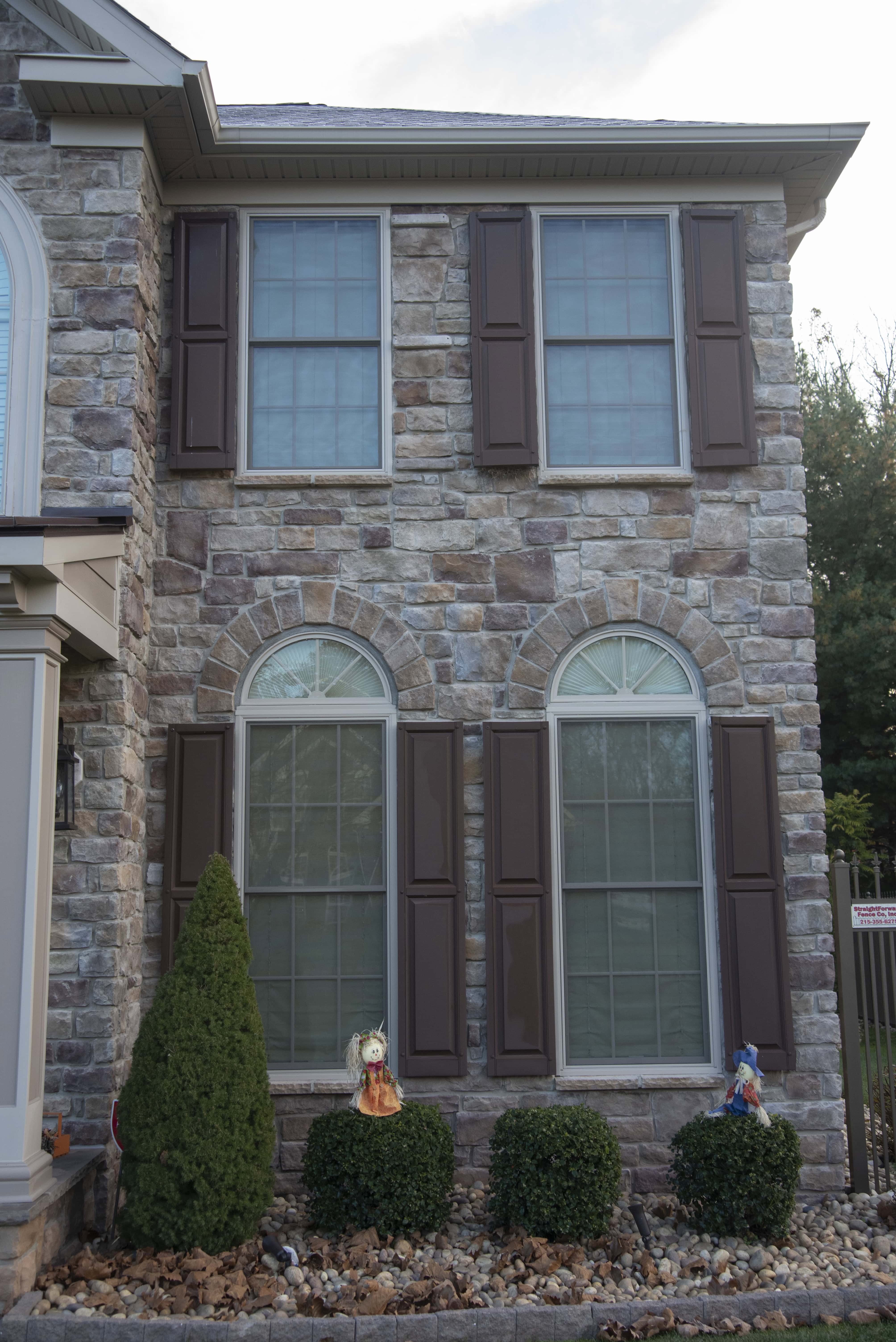 A stone façade house with two upper and two lower windows, all with closed brown shutters. Two small bushes, a conical shrub, and two decorative figurines are in the front garden.