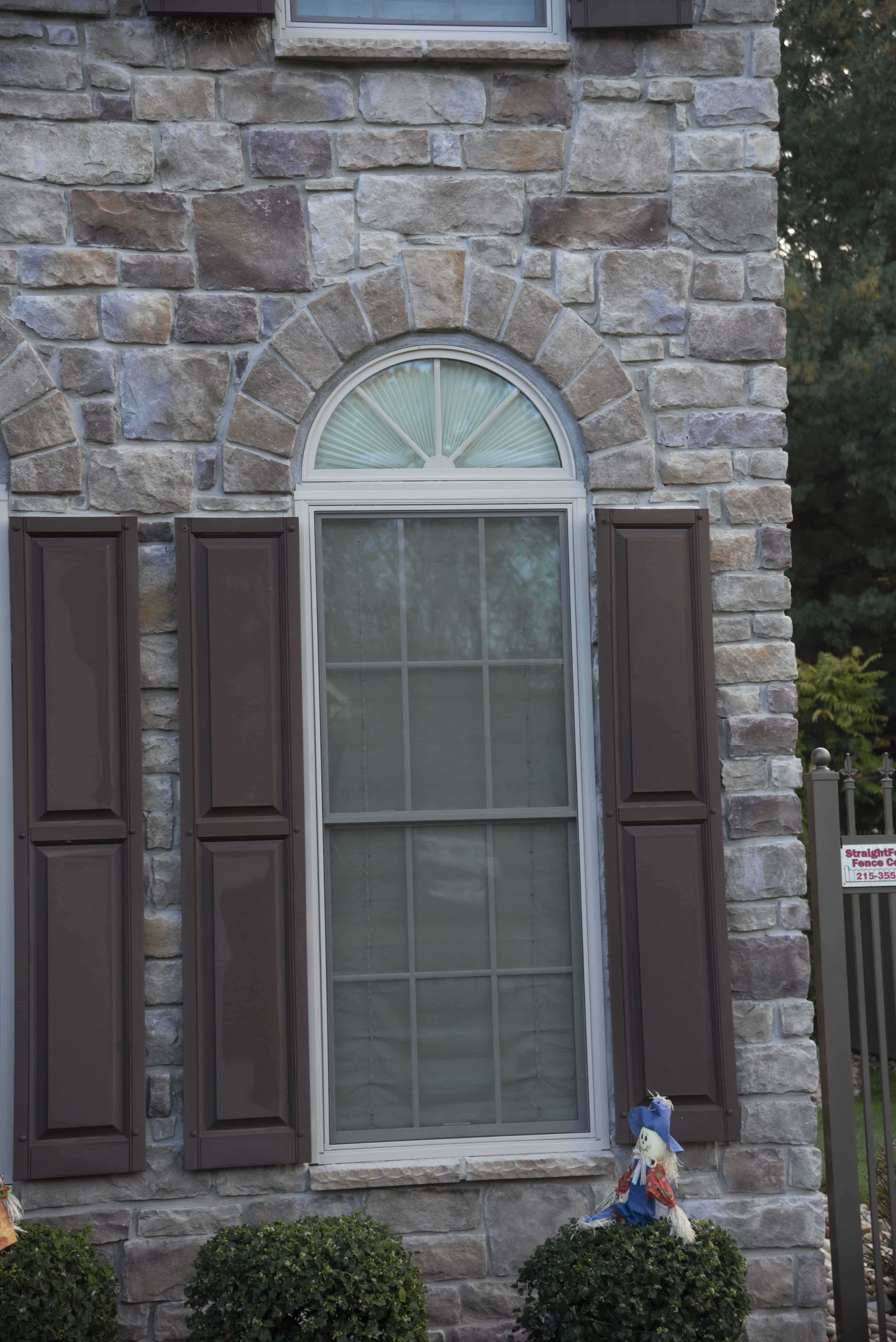 A tall window with an arched top and brown shutters on a house with stone siding. Two small bushes are planted in front of the window, and part of a decorative scarecrow is visible at the bottom right.